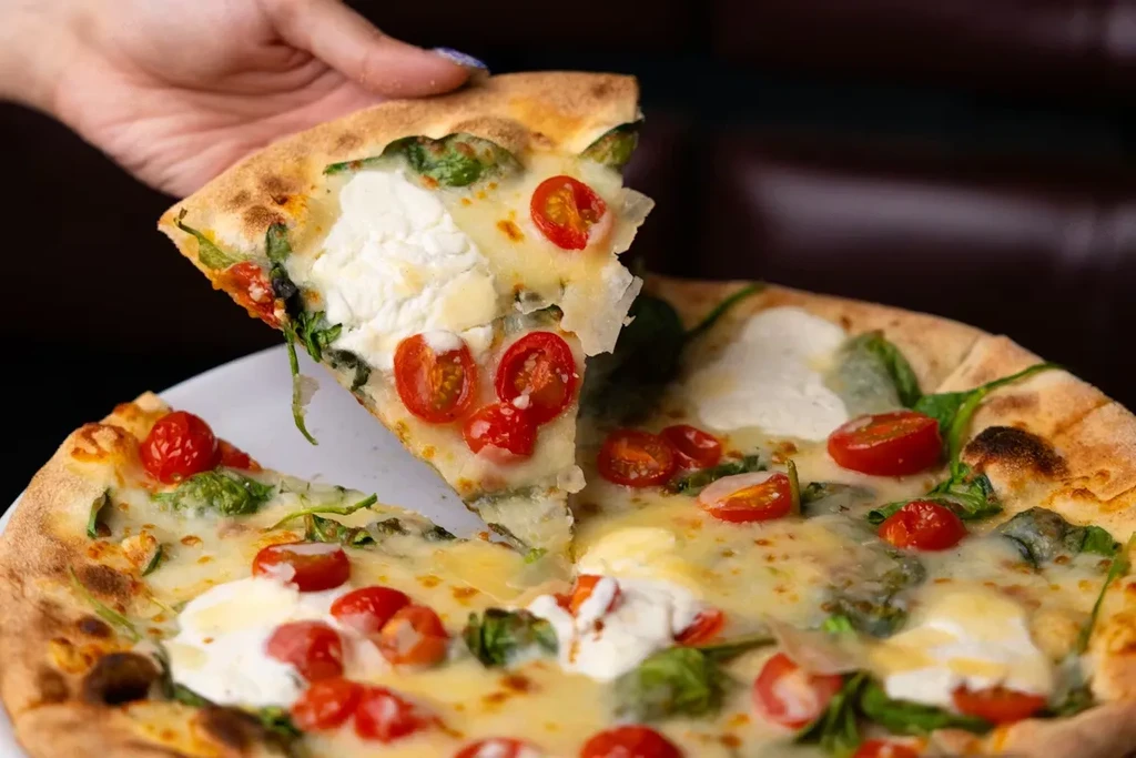 A hand lifts a pizza slice with cheese, tomatoes, greens, and ricotta.