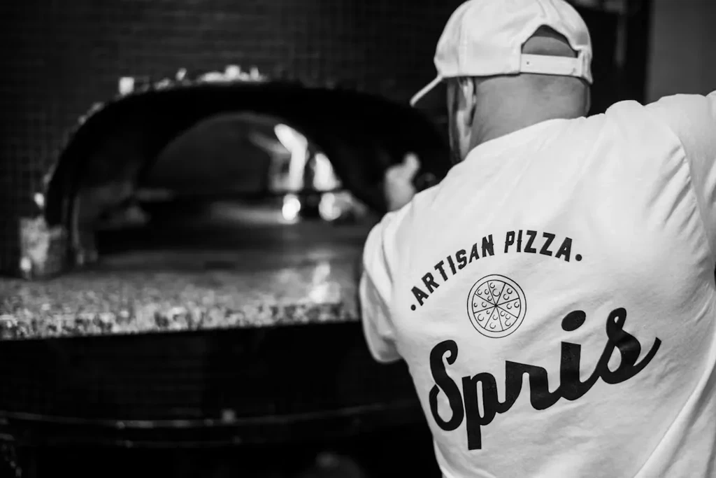 Person in "ARTISAN PIZZA" shirt faces a traditional brick pizza oven.