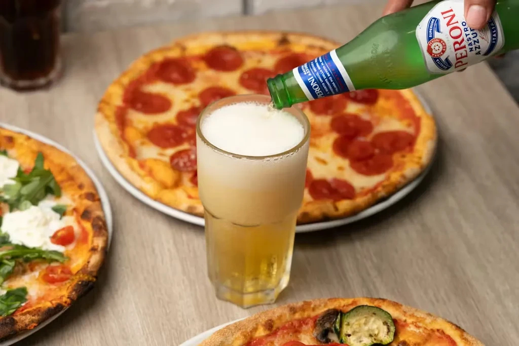 Hand pouring Peroni beer into a glass, next to a pepperoni pizza.