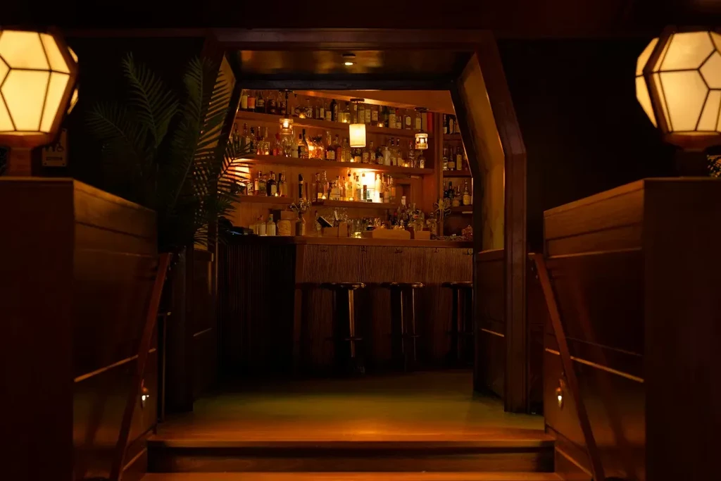 Warmly lit wooden entrance framed by geometric lanterns to a stocked bar.