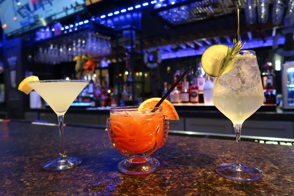 Three uniquely garnished cocktails on a dark bar with blue lights.