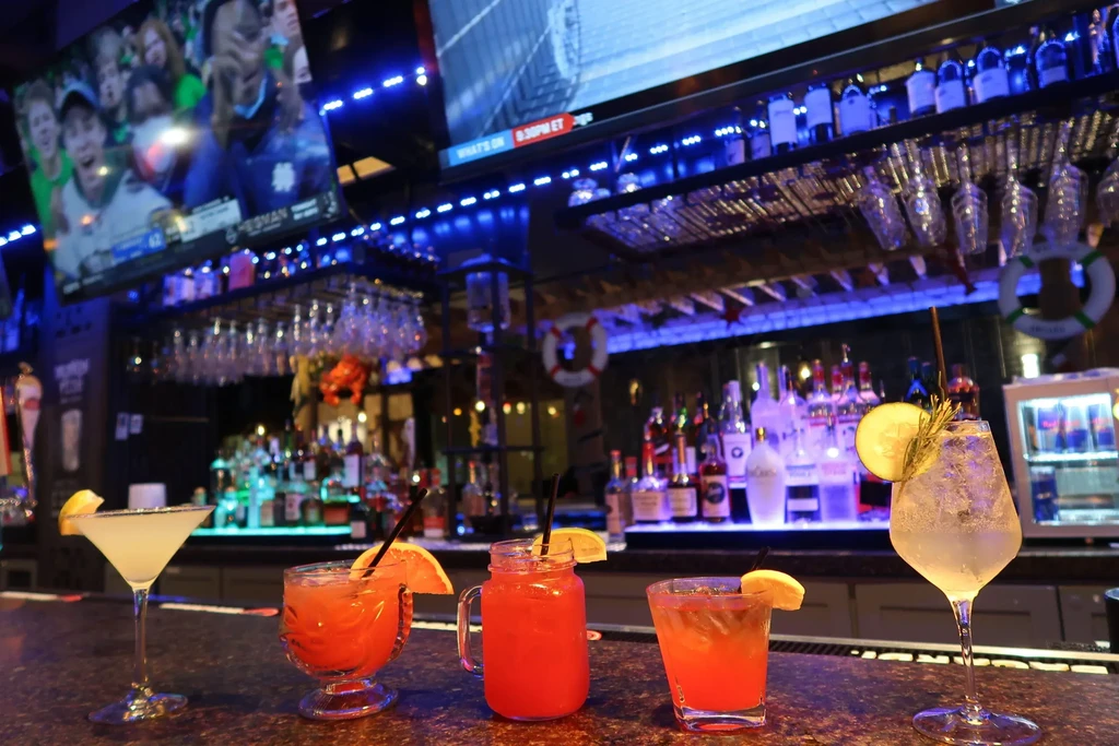 Five vibrant cocktails in front of a blue-lit bar with TVs.