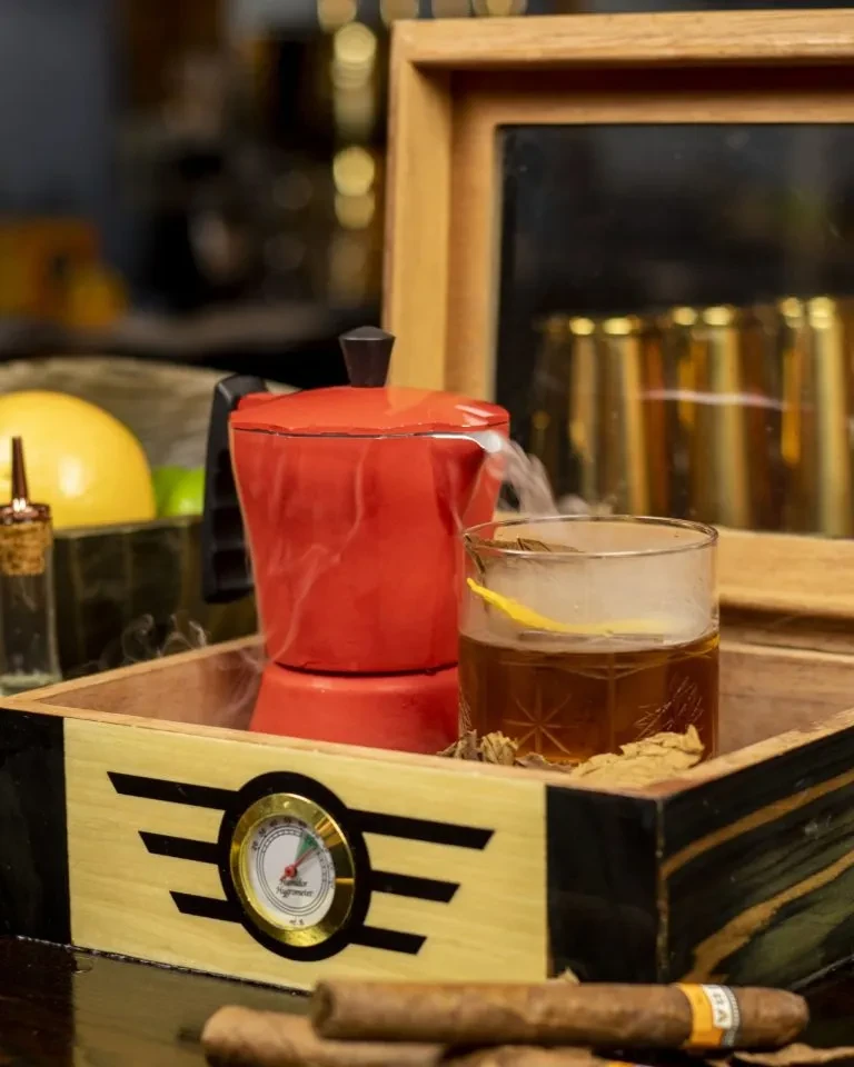 Steaming red Moka pot, amber drink with lemon, and cigars in humidor.