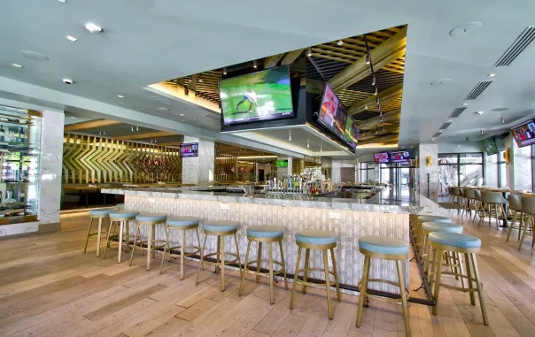 Modern bar interior with white counter, blue stools, TVs, and golden wall.