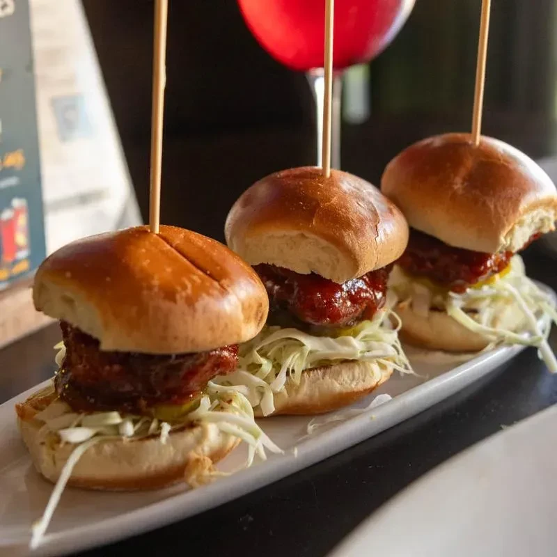 Three saucy sliders with coleslaw and pickle on a white plate.