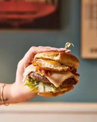 Hand holds burger with meat patty, cheese, bacon, egg, lettuce.