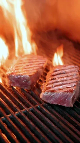 Two medium-rare steaks with grill marks cooking on a fiery grill.