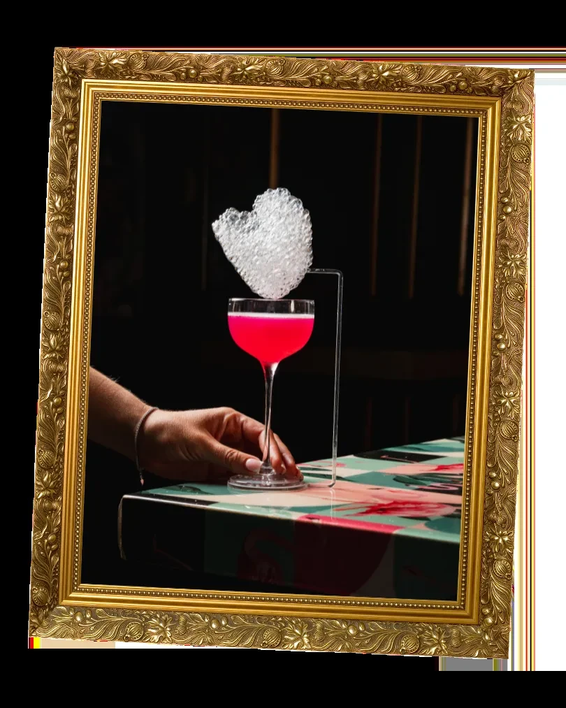 Vibrant pink cocktail with heart foam, held in an ornate gold frame.