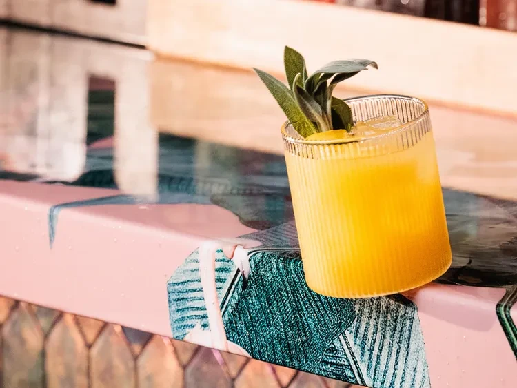 Yellow cocktail with sage and lemon on a pink patterned surface.