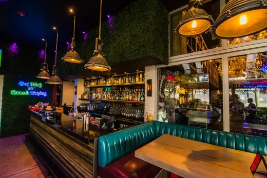 Bar with green hedge walls, liquor bottles, and 'Get Tipsy' neon sign.