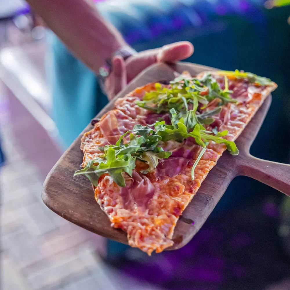 Flatbread pizza with prosciutto, arugula, and cheese on a wooden paddle.