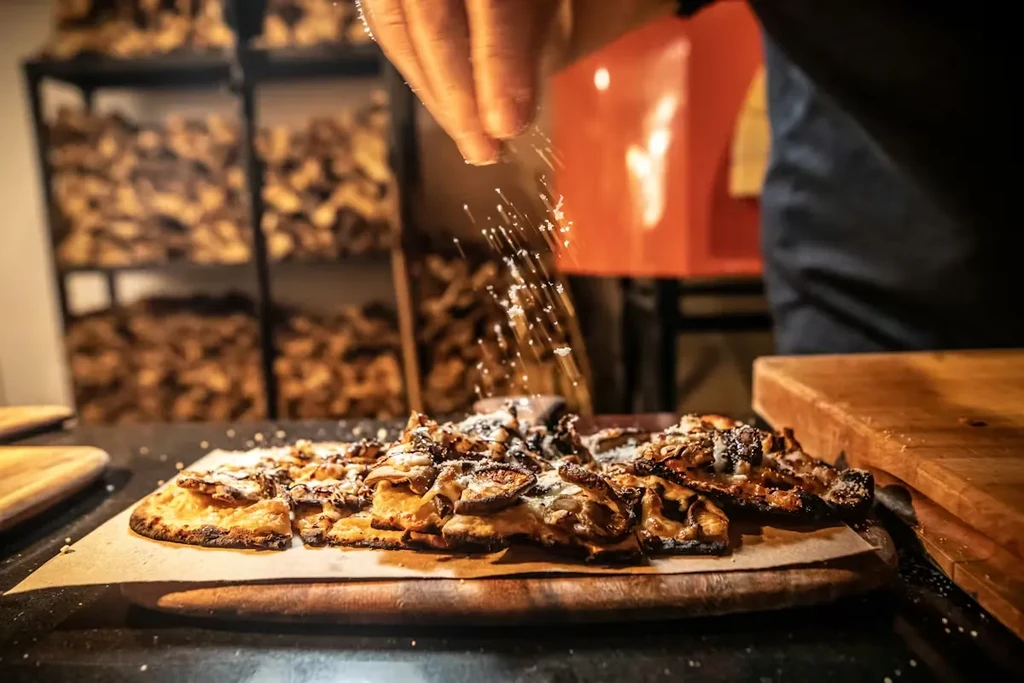 Close-up: Hand sprinkles white seasoning on rustic flatbread pizza.