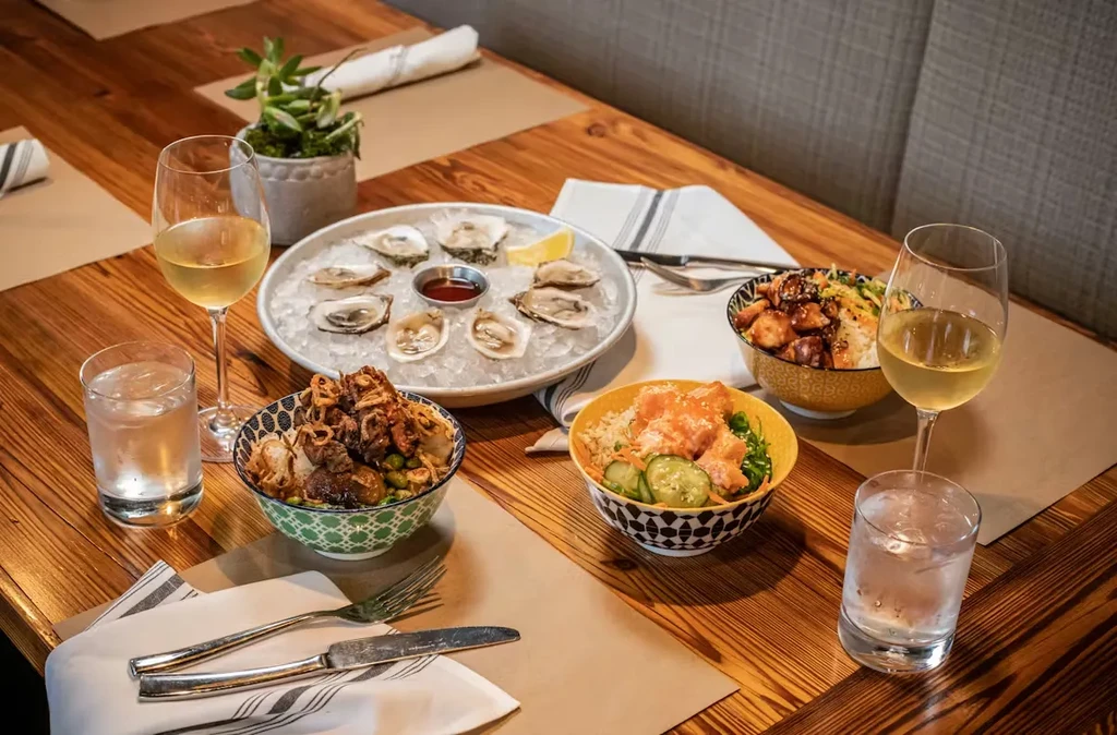 Well-set table with oysters, colorful poke bowls, wine, and water.
