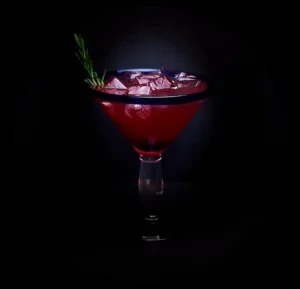 Red cocktail in martini glass with ice and rosemary against dark background.