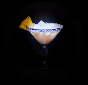 Sugar-rimmed blue cocktail glass with frothy drink, ice, and pineapple garnish.