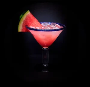 Pink cocktail with ice, blue rim, and watermelon garnish on black.