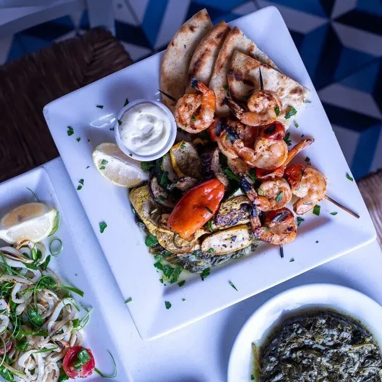 Grilled shrimp skewers, vegetables, pita, and calamari salad on patterned tiles.