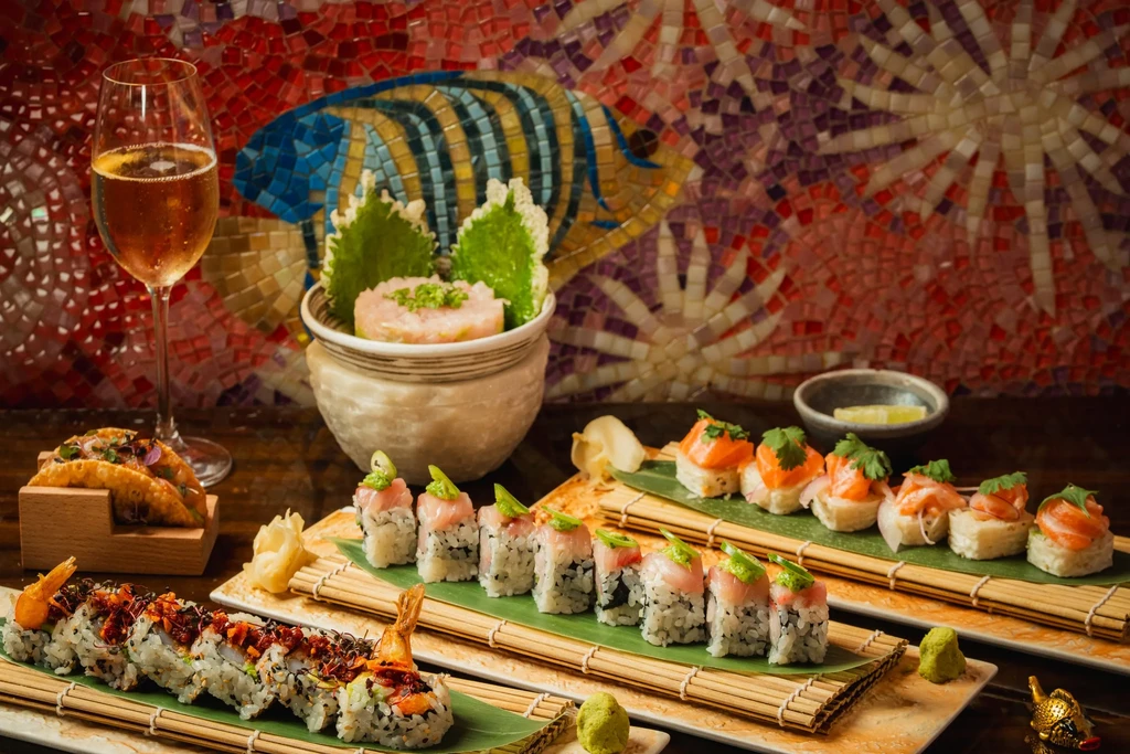 Japanese sushi, tacos, salad, and a sparkling drink, against mosaic wall.