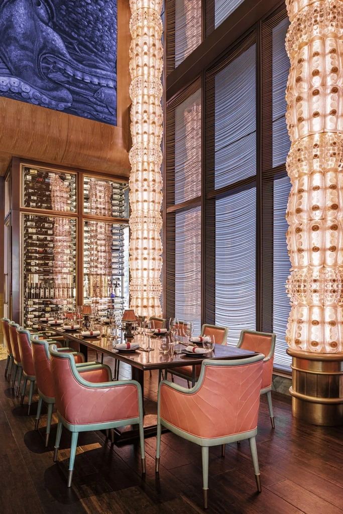 Restaurant interior: Long table, coral-teal chairs, ornate chandeliers, octopus art.