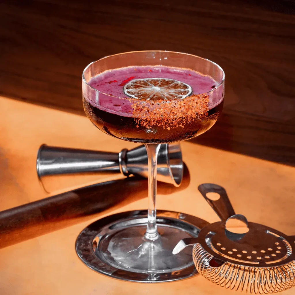 Deep red cocktail in coupe glass with spice rim and bar tools.