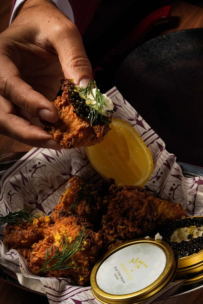 Hand holds fried chicken with caviar, cream, dill; caviar tin foreground.
