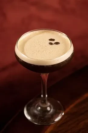 Dark cocktail in coupe glass with creamy light brown foam, coffee beans.