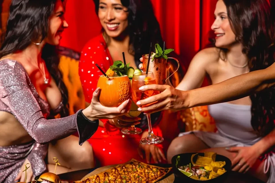 Three women toast diverse drinks and food in an upscale bar.