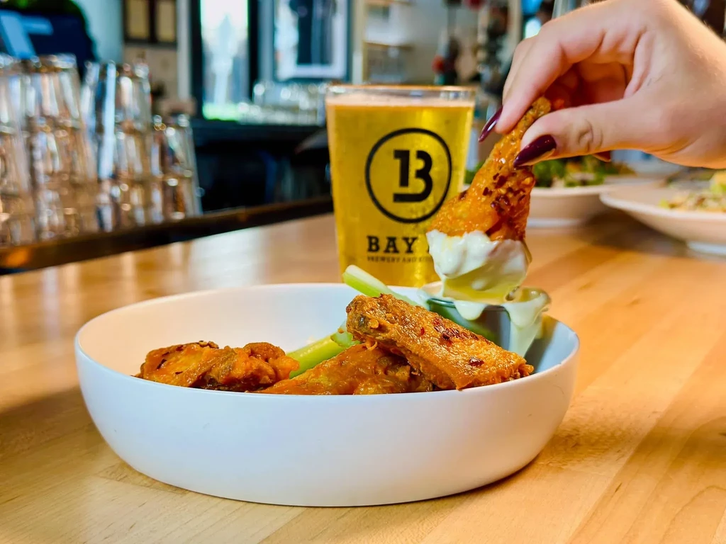 Hand dipping crispy chicken wing into sauce next to Bay Brewery beer.