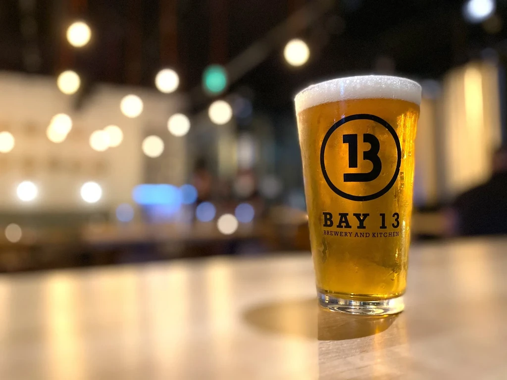 Glass of cold amber beer with a frothy head and 'BAY 13' logo.