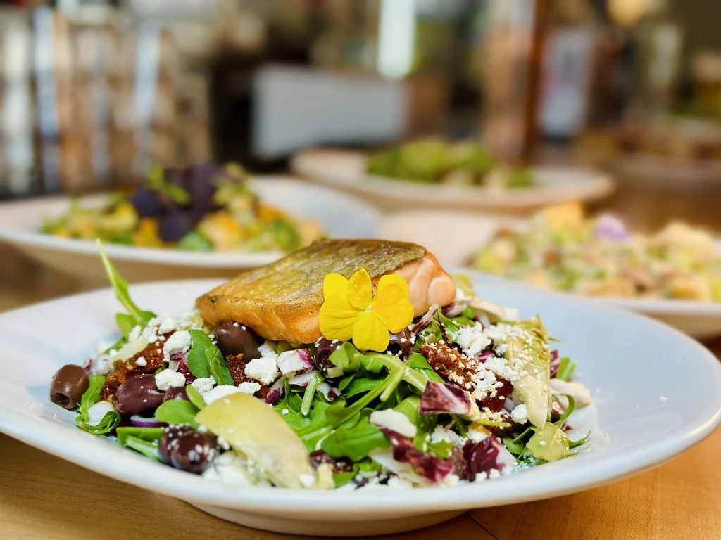 Plated grilled salmon, crispy skin, vibrant salad, feta, olives, flower.