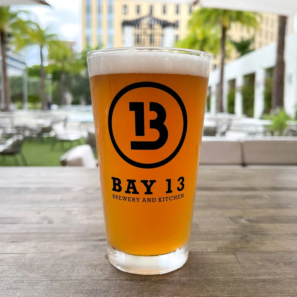 Golden beer in pint glass with Bay 13 Brewery logo outdoors.