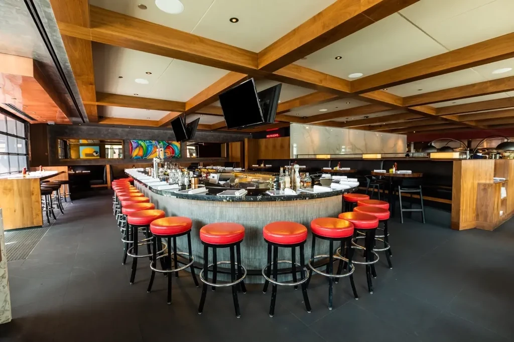 Modern bar interior with curved dark counter, red stools, wood beams.