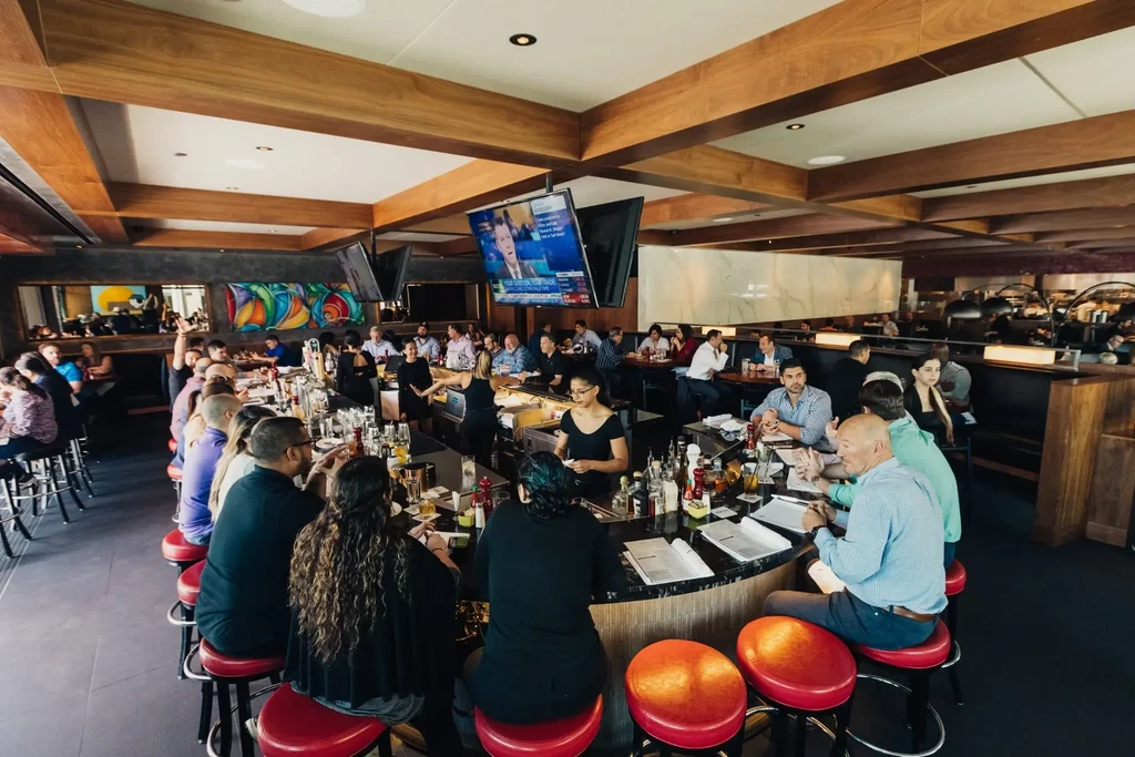 Bustling bar and restaurant interior with curved bar, patrons, and wooden beams.