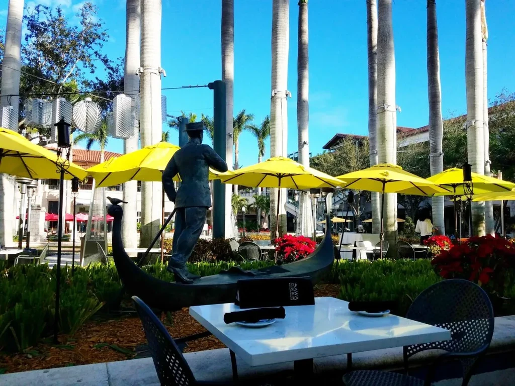Sunny outdoor plaza with yellow umbrellas, palm trees, and a gondolier statue.