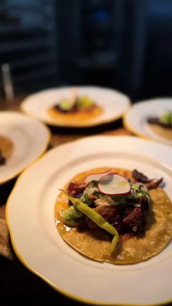 Foreground taco on white plate with meat, radish, and herbs.