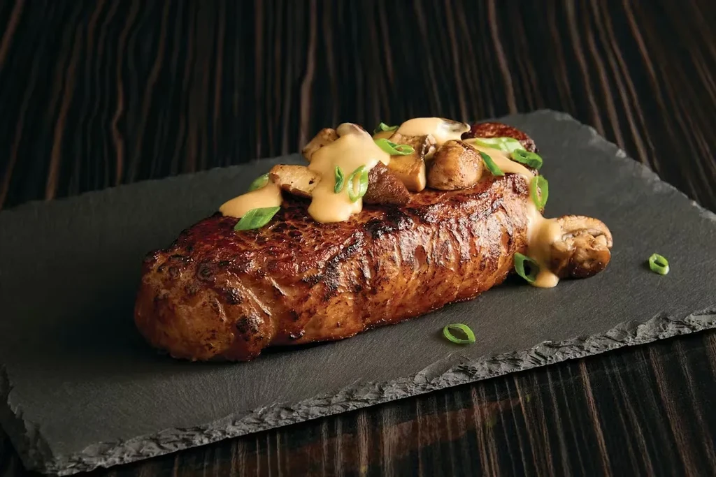 Grilled steak topped with mushrooms, creamy sauce, and herbs on a slate.