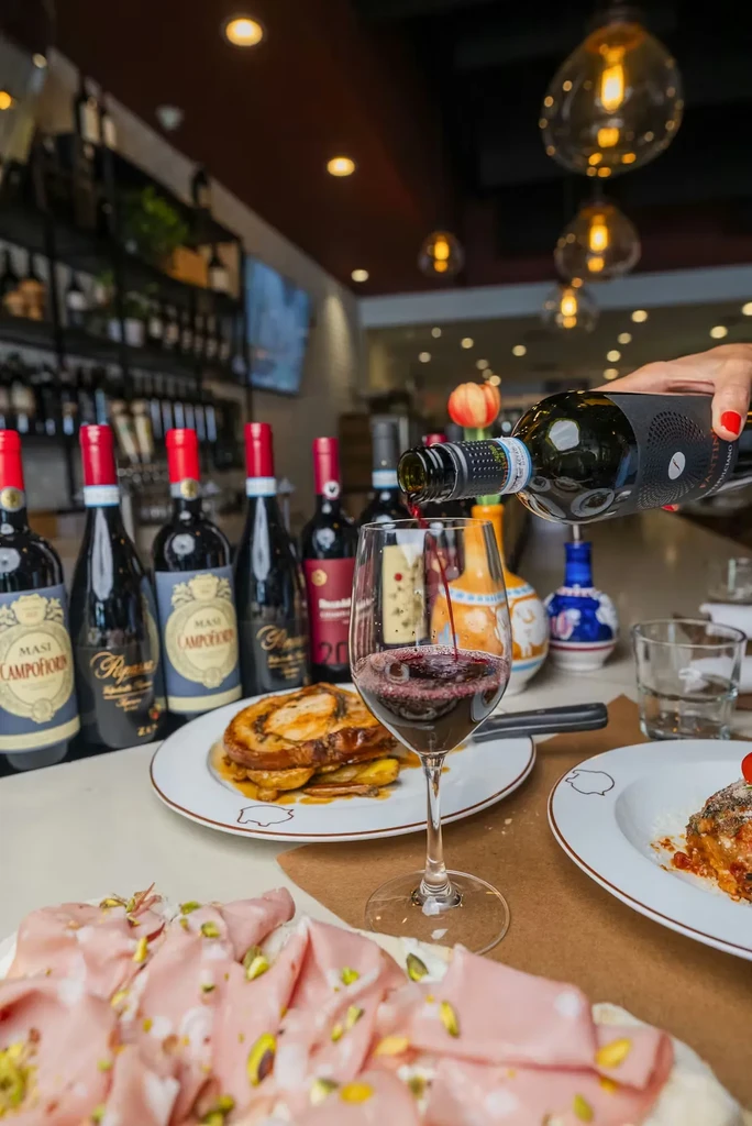 Hand pours red wine, with mortadella and various dishes on a table.