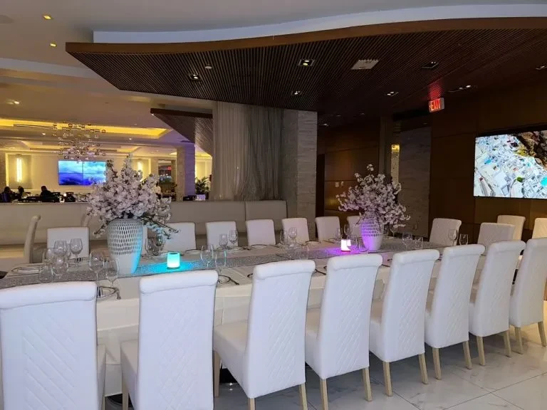 Elegant modern restaurant with long dining table, floral arrangements, and large screens.