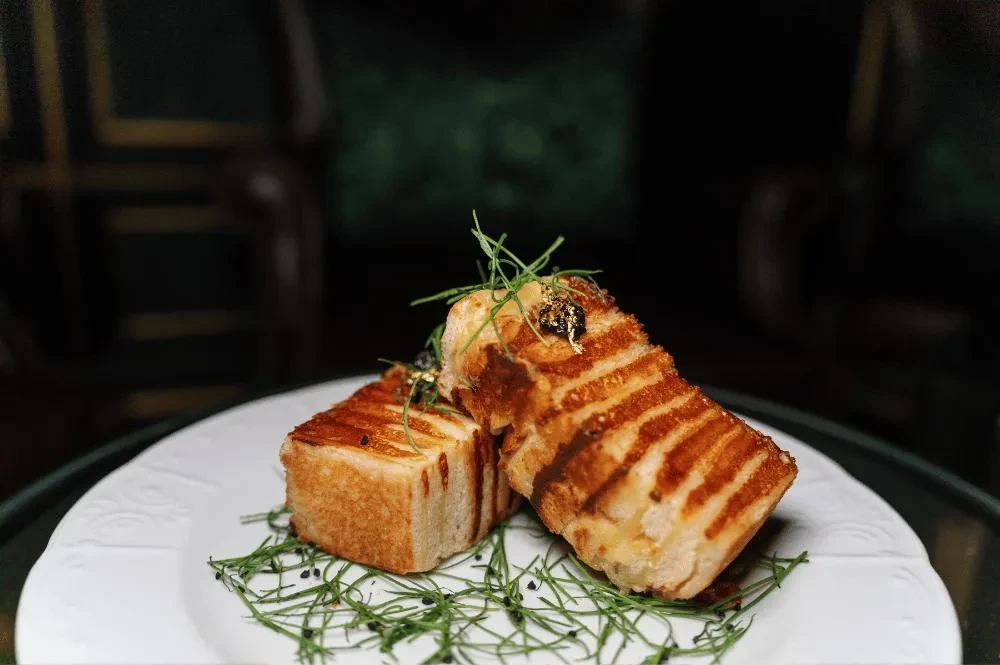Two golden-brown grilled toast pieces, garnished with herbs and gold leaf.
