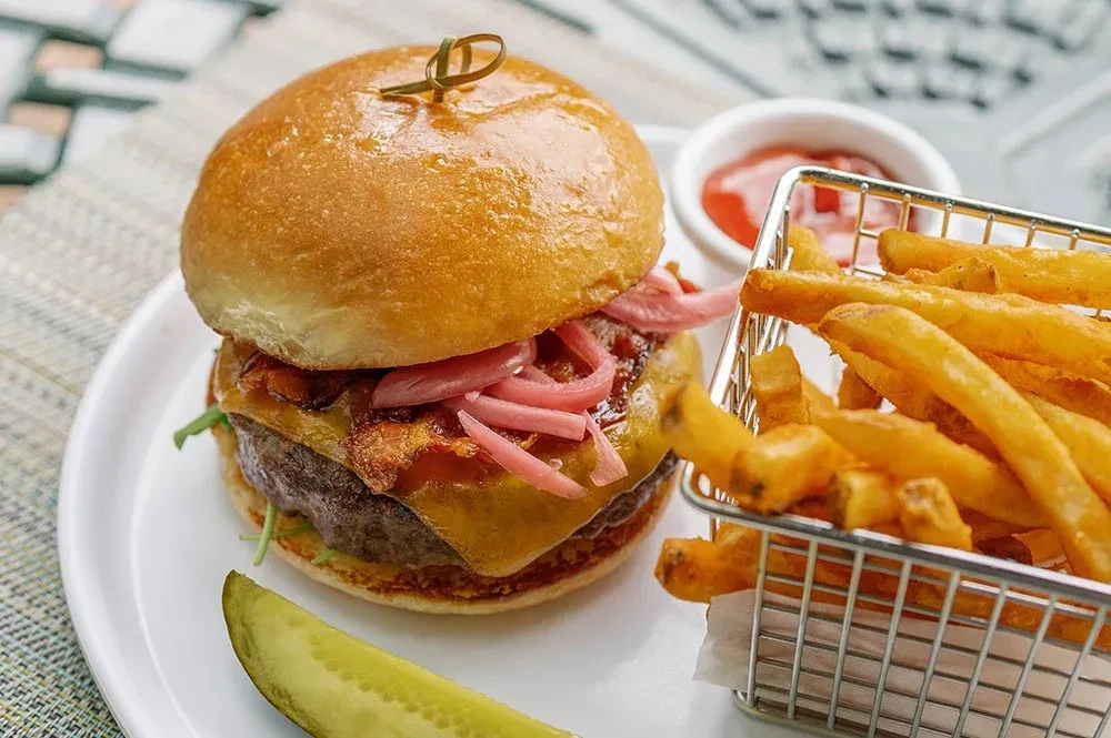 Burger with brioche bun, bacon, pink onions, fries, ketchup, and pickle.