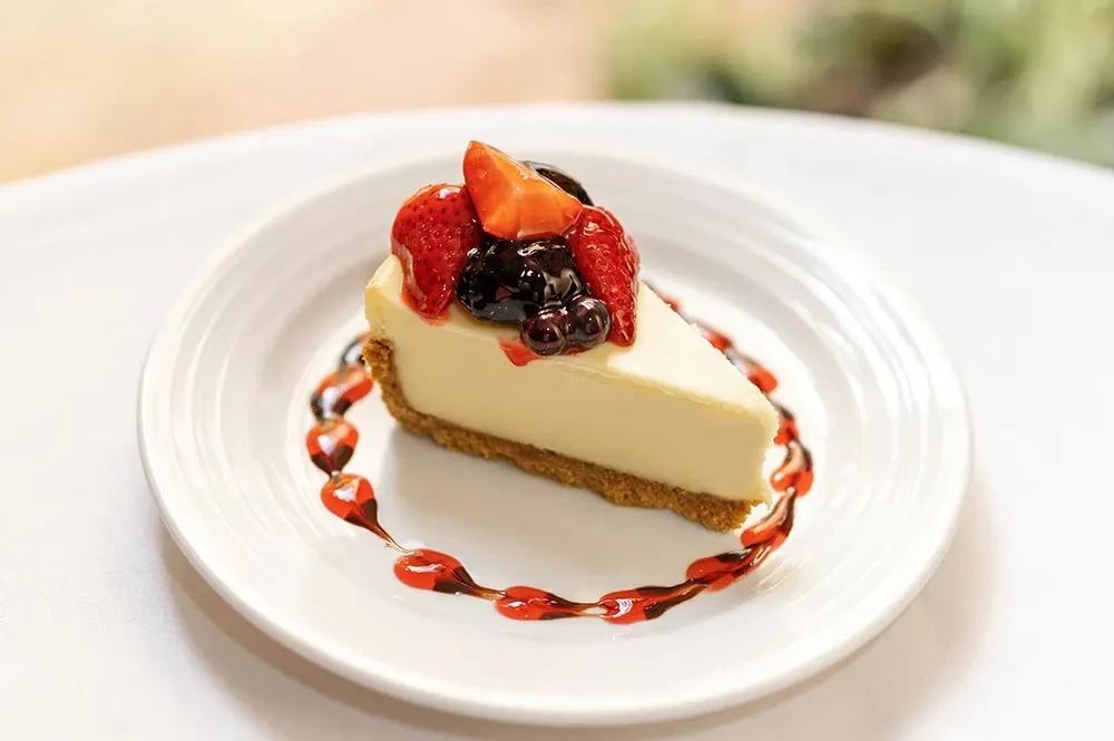 Slice of cheesecake with graham cracker crust, glazed berries, and two sauces.
