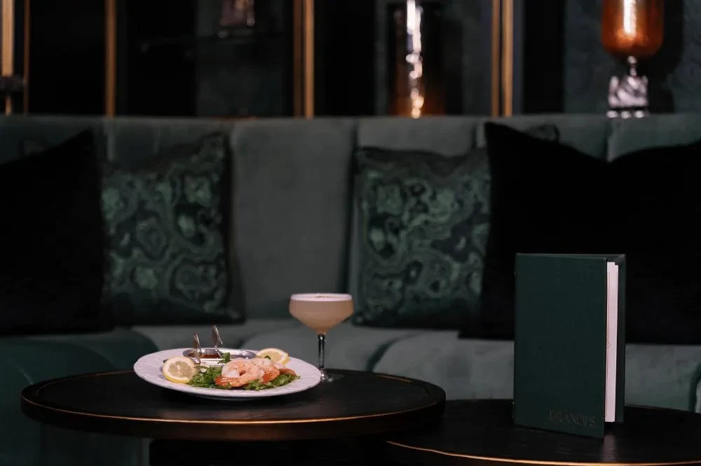 Shrimp cocktail, drink, and 'BRANDY'S' menu in a dark, plush restaurant.