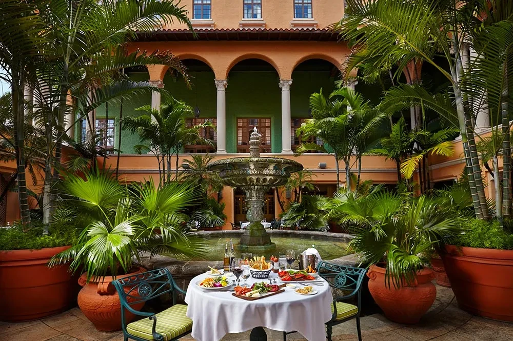 Outdoor dining table with food, fountain, and plants in an elegant courtyard.
