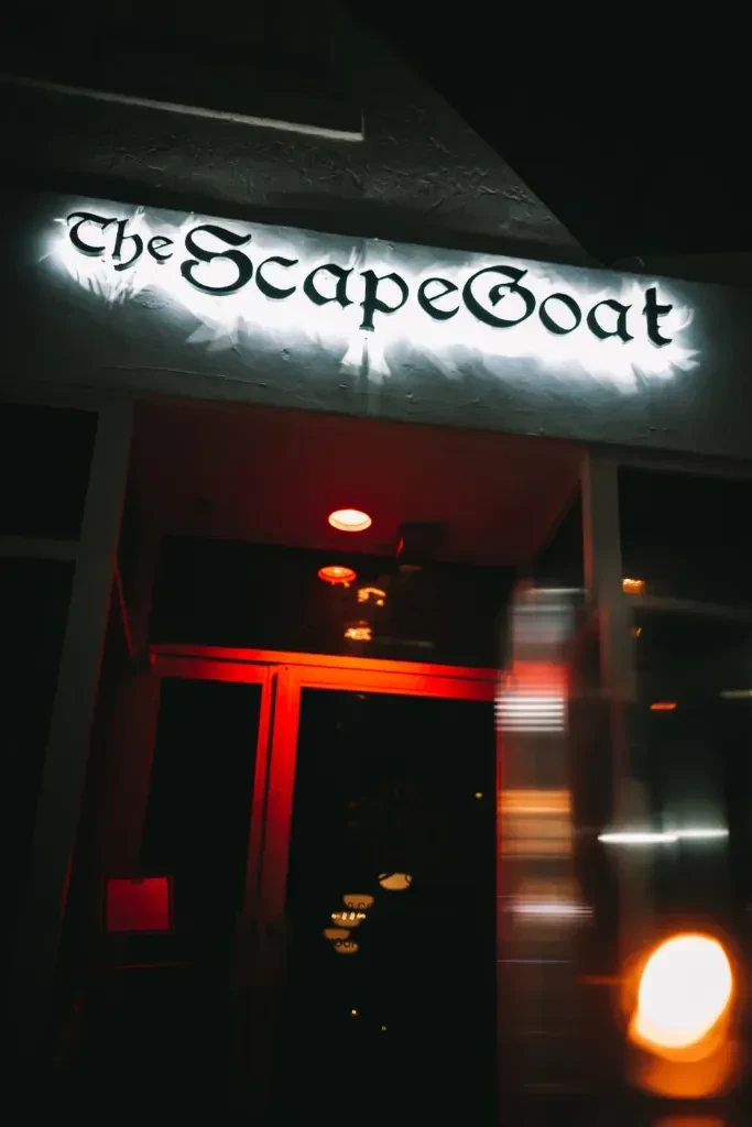 Nighttime exterior of 'The ScapeGoat' with glowing white sign and red entrance.