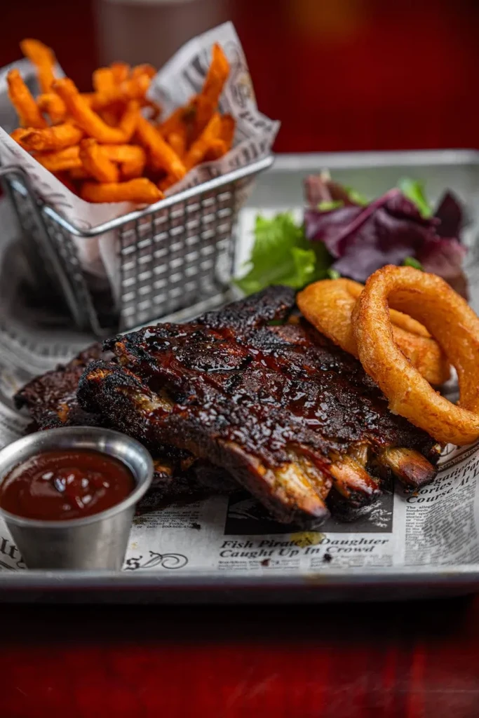 Glistening BBQ ribs, sweet potato fries, onion rings, salad on metal tray.