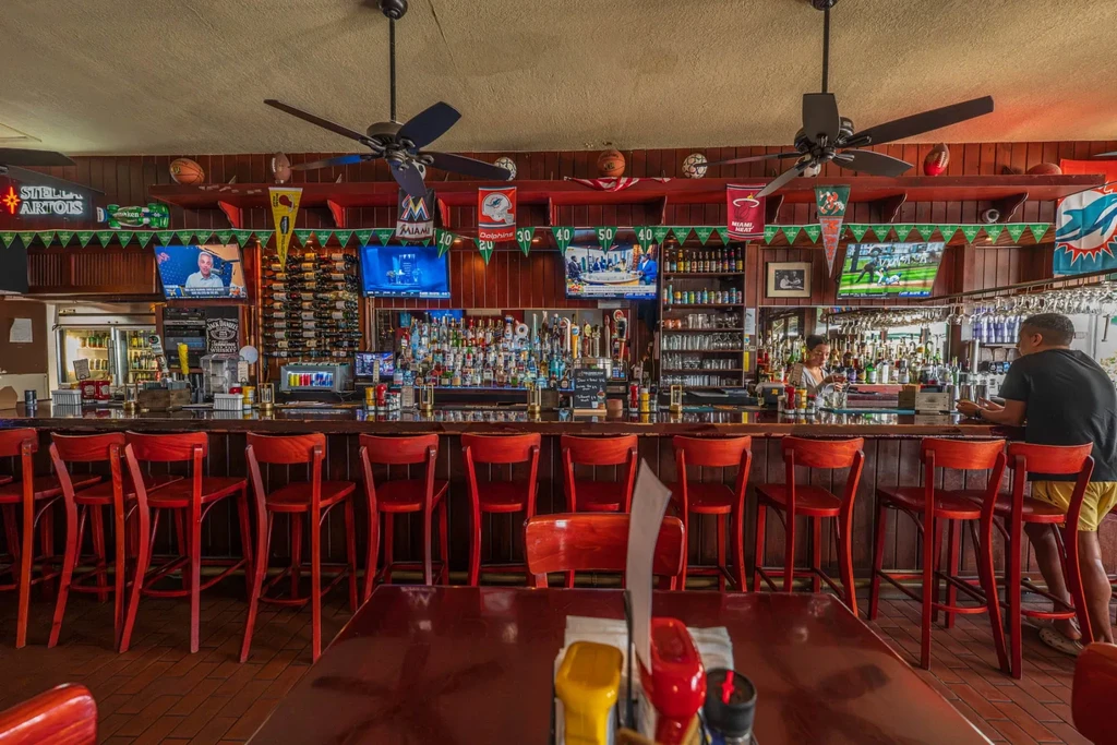 Sports bar interior with a long bar, red stools, liquor, and TVs.