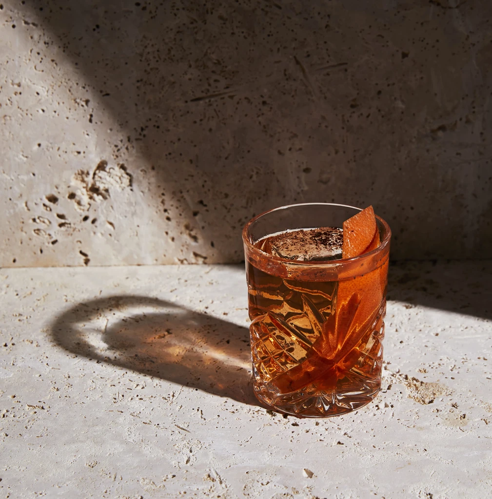 Amber cut-glass cocktail with ice, orange peel, and garnish on stone.