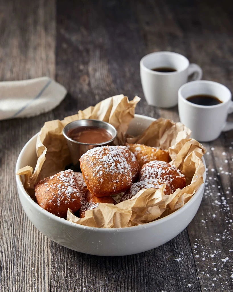 Bowl of powdered sugar beignets with chocolate sauce and coffee.