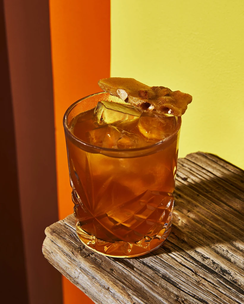 Amber cocktail with brittle garnish on rustic wood, colorful panel background.