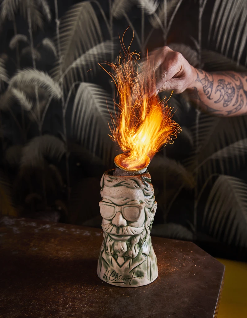 Flaming cocktail in a man-shaped tiki mug, hand visible.